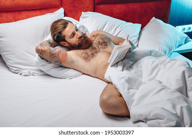 Sexy Naked Attractive Man With Mustache And Beard Lying In His White Bed And Reading A Book. Night Concept