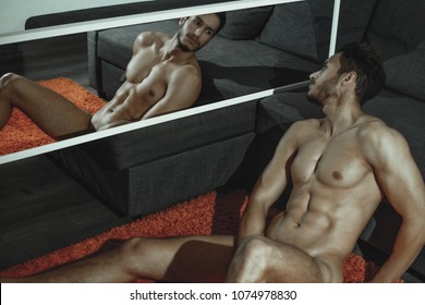 sexy and naked muscular young man posing on the floor near the sofa  and looks at her reflection in the mirror