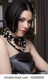 Sexy naked young caucasian adult woman with red lips, short black hair and a pierced eyebrow, covered in a dark satin sheet and wearing a black and white pearl string necklace