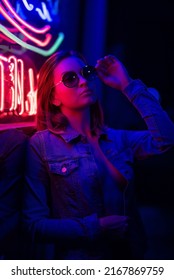 Sexy portrait of a young girl with an open bust in sunglasses and with in the night city, with creative light on the background of neon lamps.Night clubs, parties, strip business, night life