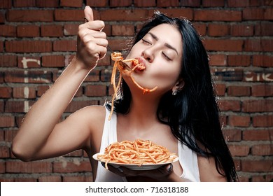 Sexy seductive cheeky girl in a white T-shirt and a plate of pasta and a fork wildly eats pasta with ketchup standing in glasses on the background of a brick wall