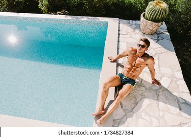 Sexy shirtless muscular man in swim trunks relax and posing near swimming pool outdoors. Muscular sports male model wearing sunglasses relax outdoors
