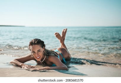 Sexy Slim Woman Girl In Bikini Is Lying On The Sand Of The Beach In The Incoming Waves Of The Sea During Vacation On Seashore, Getting Tan, Holding Feet Up. Summer Vibes On The Beach