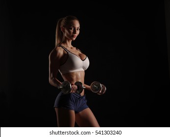 Sexy sports girl in sportswear with athletic attractive body (perfect abdominal muscles, strong biceps) is doing lifting exercises using dumbbells, fitness with equipment, dark black background