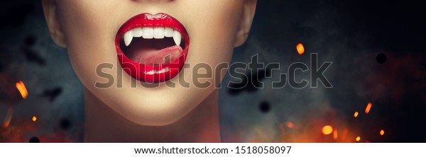 セクシーな吸血鬼女の赤い血色の唇の接写 舌で牙を舐める吸血鬼の女の子 ファッショングラマーハロウィーンのデザイン 女性の吸血鬼の口の接写 歯 黒い怖い背景にバット の写真素材 今すぐ編集