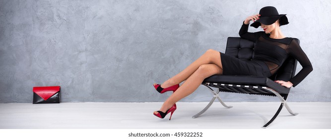 Sexy Woman In Black Dress With Hat And Red Heels Sitting On A Lounge Chair, Red-black Handbag In The Background