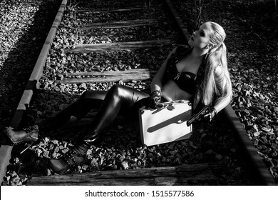 Sexy Woman In Black Leather Clothes With Gun And Suitcase. Gangsta Girl With Weapon Sitting On The Abandoned Railway At Night. 