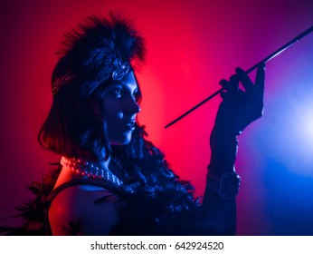 Sexy Woman Dressed In The Style Of A 20's Great Gatsby On Party. Neon Blue And Red Light. Black Dress, Pearl Beads, Beautiful Makeup And Hairstyle. Portrait Of Attractive Young Lady Smoking Mouthpiece