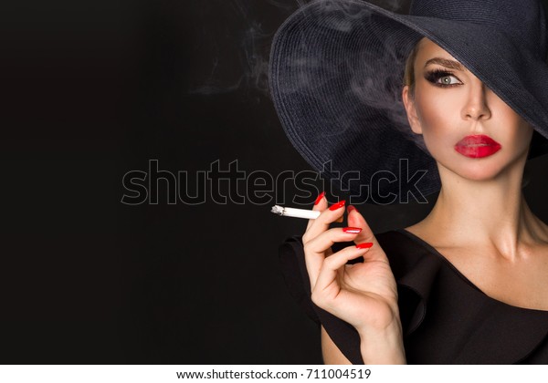 Sexy\
woman in elegant hat and with red lips blowing smoke, isolated on\
black. Femme fatale. Elegant lady with\
cigarette.