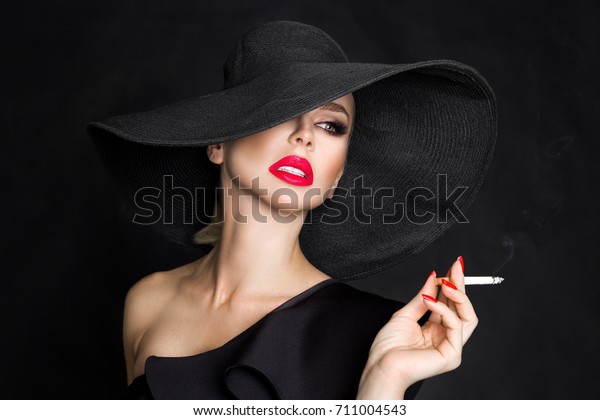 Sexy\
woman in elegant hat and with red lips blowing smoke, isolated on\
black. Femme fatale. Elegant lady with\
cigarette.