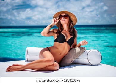 Sexy woman sitting on cozy white lounger on the beach, stylish model wearing fashionable hat and sunglasses, luxury summer vacation concept 