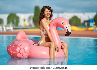 Sexy Woman Sitting On Pink Inflatable Flamingo, While Floating In Swimming Pool At Sunny Day. Happy Female In White Swimsuit Smiling, While Riding On Floaty Bird In Pool Outdoors. Concept Of Leisure.