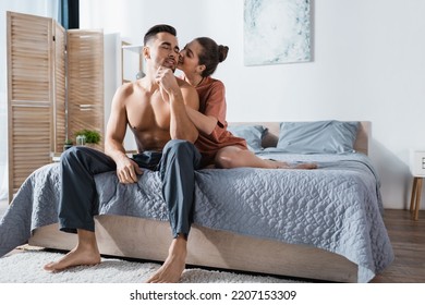 Sexy Woman In T-shirt Kissing Shirtless Man Sitting On Bed In Pajama Pants