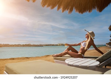 Sexy Frau mit Bikini, die unter einem Strohschirm am Strand auf einem Liegestuhl sitzt und dabei Glas mit Cocktail hält oder Getränk