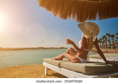 Sexy Frau mit Bikini, die unter einem Strohschirm am Strand auf einem Liegestuhl sitzt und dabei Glas mit Cocktail hält oder Getränk