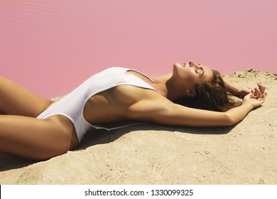 Sexy woman in white swimsuit is lying on the sand beside a lake with a pink water