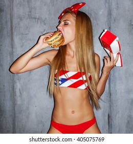 Sexy young blonde tanned sporty blonde woman eating hamburger in studio on grey background 