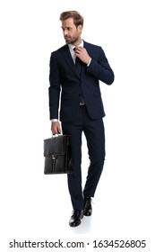 Sexy Young Businessman In Navy Blue Suit Looking To Side And Fixing Tie, Holding Suitcase And Walking Isolated On White Background, Full Body