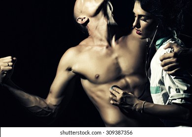 Sexy Young Couple Of Stripped Man With Strong Muscular Beautiful Body And Pretty Brunette Nurse In White Smock With Stethoscope Standing On Black Studio Background, Horizontal Picture