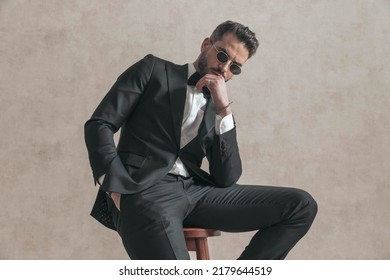 Sexy Young Guy In Elegant Black Tuxedo With Hand In Pocket Touching Lips And Being Sensual On Beige Background