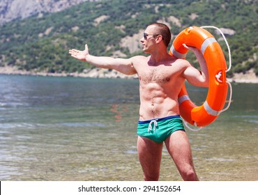 Sexy Young Man Body Builder With Six Pack Is Posing On The Beach With Orange Lifebuoy Ring