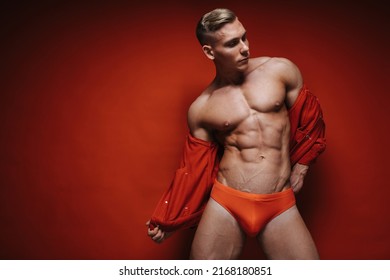 Sexy young man in red speedo and jean jacket stripping at red background. Handsome shirtless guy taking off his red jacket. Fitness male model in studio. Muscular sportsman with six pack abs.