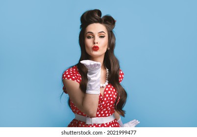 Sexy Young Pinup Woman In Old Fashioned Dress And Gloves Blowing Air Kiss At Camera Over Blue Studio Background. Portrait Of Pretty Retro Lady Being Flirty, Sending Her Love