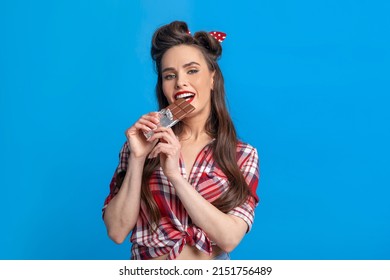 Sexy Young Pinup Woman In Retro Style Outfit Eating Chocolate Bar On Blue Studio Background. Glamorous Millennial Lady In Old Fashioned Clothes Biting Yummy Sweet Dessert