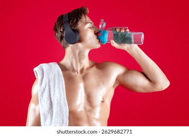 Sexy young sportsman with naked torso and towel listening to music in eadphones and drinking water on red background