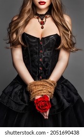 Sexy Young Woman in Black Corset Dress with Rope tied Hands holding Red Rose Flower. Art Fantasy Witch Gothic Girl Portrait with Red Lips in Historical Gown over Dark Gray Background