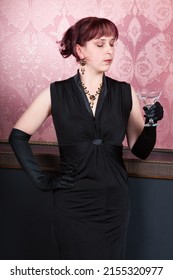 sexy young woman wearing black gown and long black gloves is standing with glass of martini in her hand