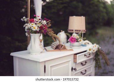 Shabby Chic Decor, White Table With Vintage Objects On It, Flowers Vase, Flower Pot An Opened Book And A Lamp, Outdoor