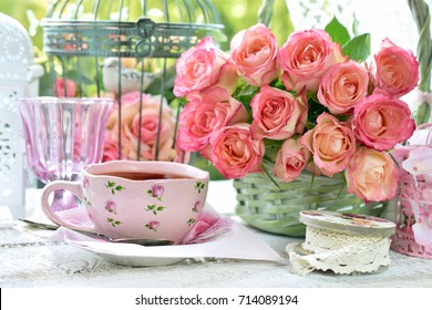 Shabby Chic Style Pink Cup Of Tea On The Table With Bunch Of Roses And Vintage Bird Cages In The Garden 