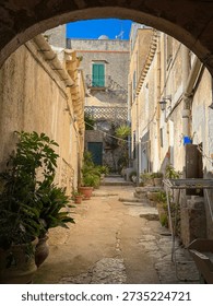 Shaded stone arch frames a small alley in Erice, Sicily, decorated with potted plants, green shutters, and sunlit walls. Intimate courtyard feel and rustic details capture charm in historic hill town.