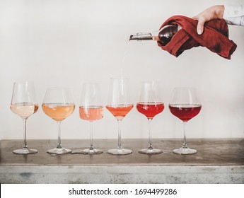 Shades Of Rose Wine In Stemmed Glasses Placed In Line From Light To Deep And Womans Hand Pouring Wine From Bottle To Glass, White Wall Background Behind. Wine Bar, Wine Shop, Tasting Concept