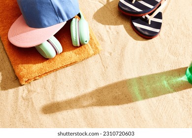 Shadow of beer bottle on warm sand with slippers, headphones, towel and cap. Summertime relaxation. Concept of alcohol drink, taste, summer vacation, holiday, brewery. Advertisement - Shutterstock ID 2310720603