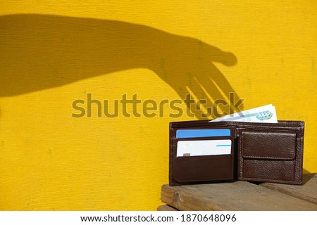 Shadow of hand on yellow wall trying to steal a leather wallet with money, banknote and credit cards. Robber pickpocket silhouette. Concept of financial crime, tax burden, unexpected expenses.