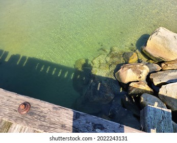 The Shadow Of A Man From Above That Is Stretched Out Atop The Water Beneath Him As The Adult Leans Over The Edge Of  Wooden Dock.