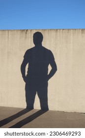 A shadow of a man on the wall. Shadow's hands are in pockets. The shadow of legs is on the floor. The wall and the floor are made of a concrete. A sunny evening sunlight in Espoo.