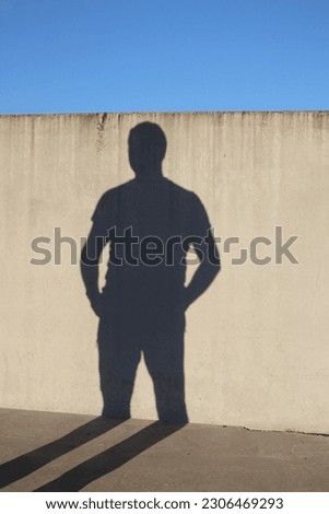 A shadow of a man on the wall. Shadow's hands are in pockets. The shadow of legs is on the floor. The wall and the floor are made of a concrete. A sunny evening sunlight in Espoo.