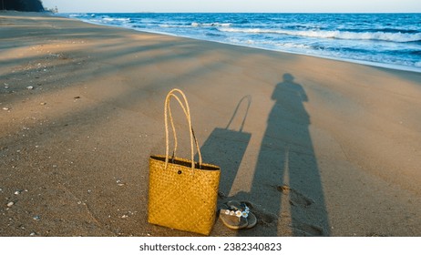 Shadow on sandy beach of a women relaxing on a lovely sunny day. Shadow of woman in beautiful light sunset at the shoreline with woven bag and sandals. - Powered by Shutterstock