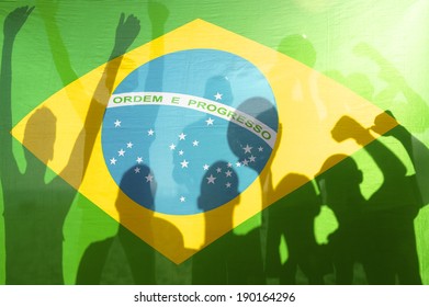 Shadow silhouettes of football team celebrating with soccer ball against Brazilian flag in bright sunlight