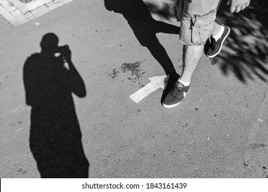 A Shadow Of A Street Photographer Sealing The Legs Of A Man Walking Against A Painted Arrow. Street Black And White Photography.