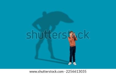 Shadow of superman or batman super hero over cool redhead guy schooler posing on blue studio wall background, making eyeglasses with fingers, copy space, collage. Childhood and imagination concept