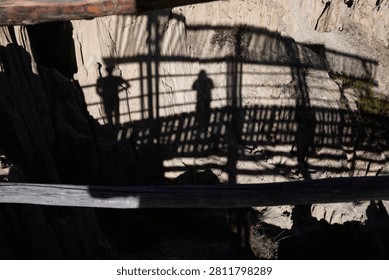 Shadow of two people standing inside a wooden bridge. The shadows are projected onto a splintered-looking mountain. Everything looks highly rustic on a sunny day.
