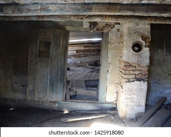 The Shadowed Interior Of An Old Abandoned Historic Timber Cabin With Sinking Uneven Floors And Walls With Brick Chimney And A Wooden Door