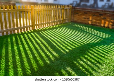 Shadows Of  A Wooden Picket Fence In A Front Yard, Front Garden With Artifical Grass As A Lawn And A Red Brick Perimiter Wall.