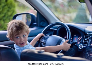 Shaggy Child Is Holding Steering Wheel In His Father's Car Like Real Driver. Boy Amusingly Plays Driver Of A Real Car. Five-year-old Boy Gets Behind The Wheel And Pretends To Drive A Car