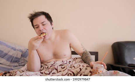 A shaggy fat man with a naked torso sits in bed, watches TV and eats pizza. The young man takes a bite of pizza and chews it, holding a milkshake in his other hand.