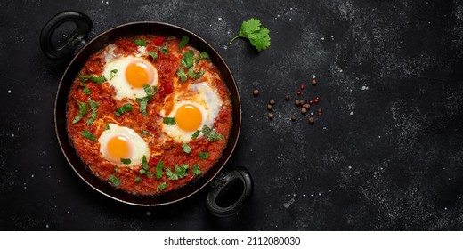 Shakshuka eggs in a pan with toast on a black concrete background. Poached eggs in a spicy tomato pepper sauce. Traditional Jewish scrambled eggs. Top view, flat lay. Banner. Textured object, selectiv